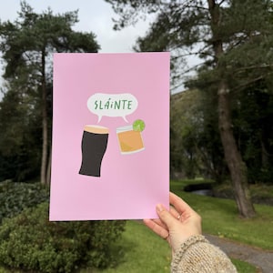 May include: A rectangular print on light pink paper featuring a dark stout beer in a glass and a golden cocktail with a lime slice. Above the drinks, a white speech bubble contains the green text SLÁINTE, representing a toast between two beverages.