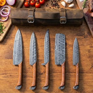 May include: A collection of five handcrafted knives with wooden handles, arranged on a wooden surface. A brown leather knife roll is visible, along with a cutting board with sliced vegetables and a steak. The knives have textured blades.