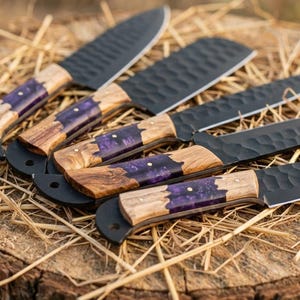 K&ouml;nnte beinhalten: Eine Auswahl handgefertigter Messer mit schwarzen Klingen und Holzgriffen, die mit lila Harz eingelegt sind. Die Messer liegen auf einer rustikalen Holzoberfl&auml;che.