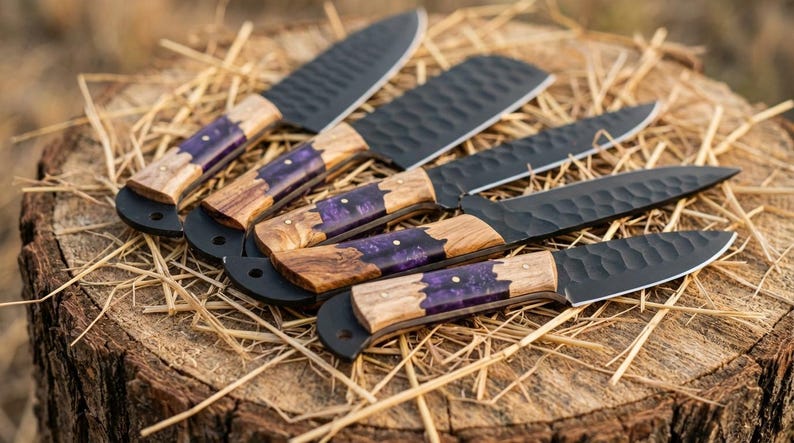 K&ouml;nnte beinhalten: Eine Auswahl handgefertigter Messer mit schwarzen Klingen und Holzgriffen, die mit lila Harz eingelegt sind. Die Messer liegen auf einer rustikalen Holzoberfl&auml;che.