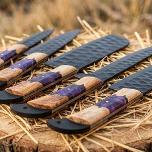 K&ouml;nnte beinhalten: F&uuml;nf handgefertigte Messer mit schwarzen Klingen und Holzgriffen. Die Griffe haben eine violette Einlage. Die Messer liegen auf Stroh, das auf einem Baumstumpf platziert ist. Die Klingen haben eine strukturierte Oberfl&auml;che.