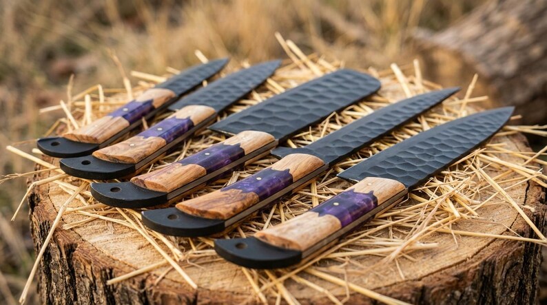 K&ouml;nnte beinhalten: F&uuml;nf handgefertigte Messer mit schwarzen Klingen und Holzgriffen. Die Griffe haben eine violette Einlage. Die Messer liegen auf Stroh, das auf einem Baumstumpf platziert ist. Die Klingen haben eine strukturierte Oberfl&auml;che.