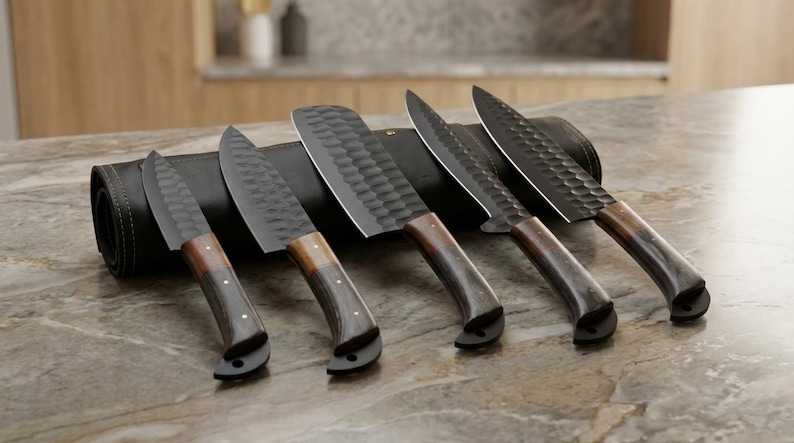 May include: A set of five kitchen knives with dark blades and wooden handles, displayed on a countertop. The blades have a textured pattern, and the handles transition from light to dark brown. A black leather knife roll is behind the knives.