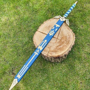 May include: A blue and gold decorative sword with a detailed design, resting on a wooden stump against a backdrop of green grass. The sword features a blue blade with gold accents and a gold hilt. The handle is wrapped in a light blue and gold pattern.