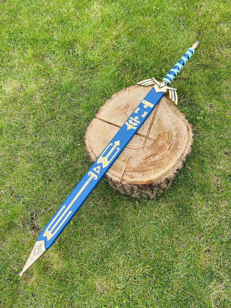May include: A blue and gold decorative sword with a detailed design, resting on a wooden stump against a backdrop of green grass. The sword features a blue blade with gold accents and a gold hilt. The handle is wrapped in a light blue and gold pattern.