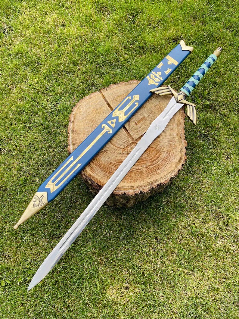 May include: A silver sword with a gold hilt and a blue and gold scabbard. The sword is resting on a wooden surface. The scabbard is blue with gold accents and a gold tip. The handle of the sword is wrapped in blue and green.
