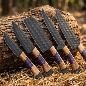K&ouml;nnte beinhalten: Eine Sammlung von f&uuml;nf handgefertigten K&uuml;chenmessern mit dunklen, strukturierten Klingen und Holzgriffen. Die Griffe haben einen violetten Akzent in der N&auml;he der Klinge. Die Messer werden auf einem Baumstamm pr&auml;sentiert.