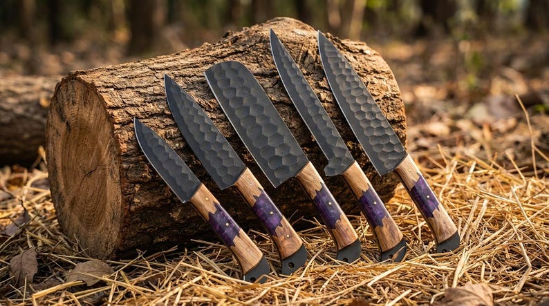 K&ouml;nnte beinhalten: Eine Sammlung von f&uuml;nf handgefertigten K&uuml;chenmessern mit dunklen, strukturierten Klingen und Holzgriffen. Die Griffe haben einen violetten Akzent in der N&auml;he der Klinge. Die Messer werden auf einem Baumstamm pr&auml;sentiert.