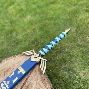 May include: A blue and gold decorative sword with a wrapped handle. The sword features a blue blade with gold accents and a gold hilt. The handle is wrapped in blue and light blue material. The sword is resting on a wooden surface.