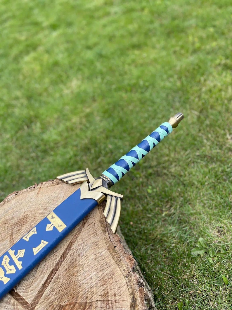 May include: A blue and gold decorative sword with a wrapped handle. The sword features a blue blade with gold accents and a gold hilt. The handle is wrapped in blue and light blue material. The sword is resting on a wooden surface.