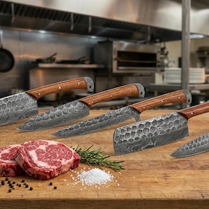 May include: A set of five kitchen knives with patterned steel blades and wooden handles. The knives are displayed on a wooden surface with two raw steaks, rosemary, salt, and peppercorns. The background is a commercial kitchen.