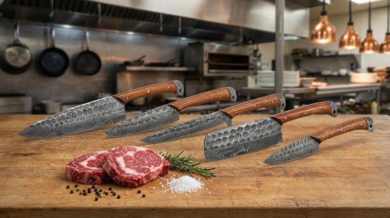 May include: A set of five kitchen knives with patterned steel blades and wooden handles. The knives are displayed on a wooden surface with two raw steaks, rosemary, salt, and peppercorns. The background is a commercial kitchen.