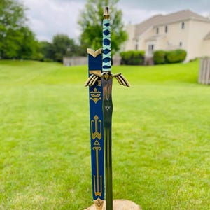 May include: A decorative sword with a blue and gold sheath, standing upright on a tree stump. The sword features a silver blade, a blue and white handle, and gold accents. The sheath is dark blue with gold detailing.