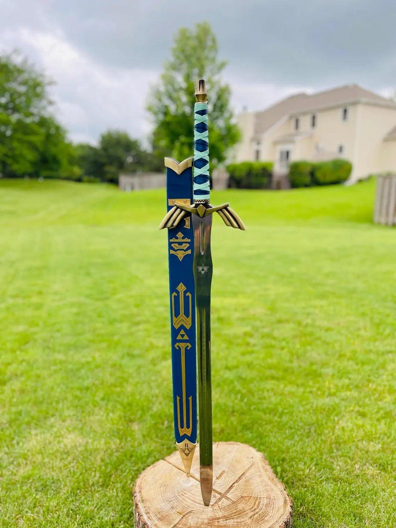 May include: A decorative sword with a blue and gold sheath, standing upright on a tree stump. The sword features a silver blade, a blue and white handle, and gold accents. The sheath is dark blue with gold detailing.