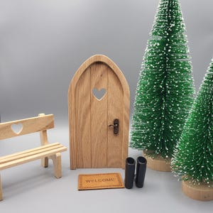 Könnte beinhalten: Eine Miniatur-Feentür aus Holz mit herzförmigem Ausschnitt und einer kleinen Fußmatte mit der Aufschrift „Willkommen“. Zwei kleine grüne Weihnachtsbäume und eine Holzbank sind ebenfalls vorhanden. Zwei schwarze Stiefel stehen vor der Tür.