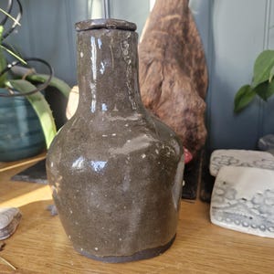 Puede incluir: Un jarrón de cerámica gris oscuro con tapa. El jarrón tiene un cuerpo redondeado y un cuello estrecho. La superficie tiene un aspecto moteado. El jarrón está sobre una superficie de madera.