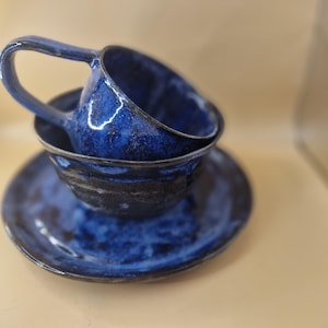 May include: A handmade ceramic teacup and saucer set. The set features a deep blue glaze with a textured appearance. The teacup is stacked on top of the saucer, with the handle visible. The set is designed for serving tea or coffee.