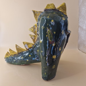 May include: A handmade ceramic dinosaur sculpture, glazed in dark green with yellow detailing. The sculpture features a head and neck, with yellow spikes running along the back. The piece has a glossy finish.
