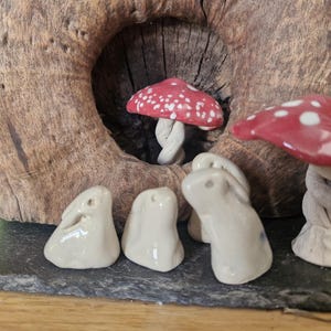 May include: A charming arrangement of ceramic mushrooms with red and white spotted caps, and small white ceramic bunny figures. The mushrooms have twisted stems. The scene is set against a backdrop of a wooden log with a natural hole.