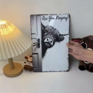 May include: A rectangular metal sign with a humorous design. It features a Highland cow looking from behind a door, with the text "Are You Pooping?" above. The cow is in black and white, giving a vintage feel to the sign.