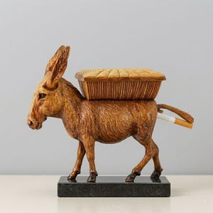 May include: A detailed wooden donkey sculpture with a basket on its back and a cigarette in its tail. The donkey is brown and stands on a black rectangular base. The basket is a lighter brown with a woven texture.