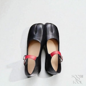 May include: A pair of black leather Mary Jane style shoes with a split toe design. The shoes have a red ankle strap with a silver buckle. The interior of the shoe is a light beige color. The shoes are on a white background.