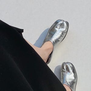 Peut inclure: Chaussures argentées métalliques à bout fendu. Les chaussures ont un aspect texturé et une semelle noire. Les chaussures sont portées avec un pantalon noir. Le design est une version moderne d'une chaussure tabi japonaise traditionnelle.