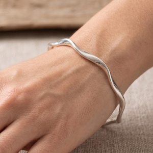 Pode incluir: Uma pulseira rígida de prata polida com um design ondulado contínuo, suave e fluido. O metal tem um acabamento brilhante e reflexivo que capta a luz, criando reflexos subtis ao longo da forma orgânica e curva da banda da pulseira.