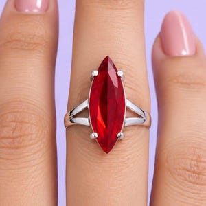 Può includere: Un anello color argento con una grande pietra preziosa rosso intenso dal taglio marquise, fissata da una montatura a quattro griffe. Il gambo dell'anello si divide in due sottili bracci metallici color argento lucido che si uniscono alla base.