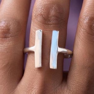 Könnte beinhalten: Ein silberfarbener offener Ring mit zwei vertikalen, rechteckigen Stäben, die sich mit einer Lücke in der Mitte gegenüberstehen. Das Band ist glatt und poliert und erzeugt einen klaren, metallischen Look, der den Finger mit einem modernen Design umschließt.