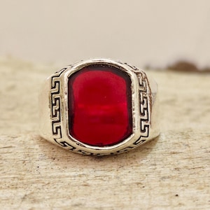 May include: A silver-toned ring featuring a large, rectangular, deep red stone set in the center. The metal band is decorated with a black-accented Greek key pattern that frames the stone, creating a bold and structured appearance.