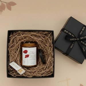 Puede incluir: Un juego de regalo con una vela en un tarro de cristal con la etiqueta "Ethan" e ilustraciones de amapolas rojas. La vela está en una caja negra con papel triturado, una caja de cerillas y una caja de regalo negra con una cinta "JUST FOR YOU".