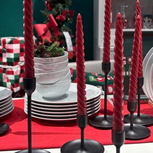 Puede incluir: Exhibición de velas rojas en espiral en candelabros de metal negro. Las velas son retorcidas y de un color rojo intenso. Los candelabros tienen un diseño sencillo y elegante. La escena está sobre un mantel rojo, con decoraciones navideñas al fondo.