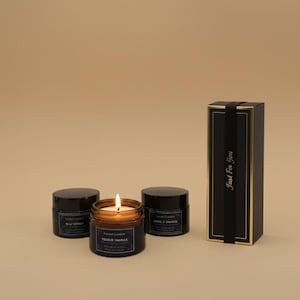 Peut inclure: Trois bougies parfumées dans des pots en verre avec des couvercles noirs. Une bougie est allumée, et l'étiquette indique "Velour Vanille". Les autres étiquettes indiquent "Bleu Voyage" et "Soleil D'Orange". Un coffret cadeau noir avec des bordures dorées et le texte "Just For You" est également présent.