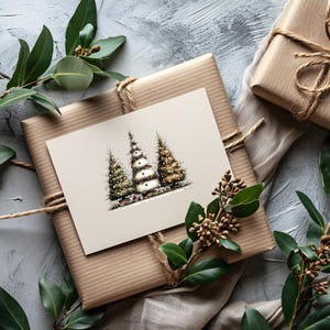 May include: A rectangular greeting card featuring a detailed illustration of three stylized Christmas trees in green, white, and gold tones. The card is placed on a square gift box wrapped in brown paper and tied with natural twine string.
