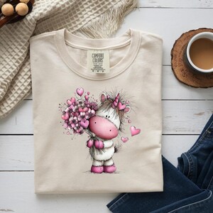 Puede incluir: Una camiseta beige Comfort Colors con un unicornio de dibujos animados sosteniendo un ramo de flores rosas en forma de coraz&oacute;n. El unicornio tiene una nariz, pezu&ntilde;as y un cuerno rosas. Corazones rosas flotan alrededor del unicornio. La camisa est&aacute; doblada sobre una superficie blanca.