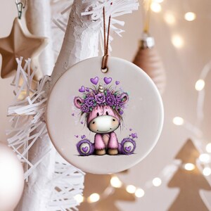 Puede incluir: Adorno navide&ntilde;o redondo de cer&aacute;mica con un unicornio de dibujos animados con una corona floral morada y detalles de coraz&oacute;n. El unicornio es rosa con una cara blanca y est&aacute; sentado. El adorno cuelga de una cuerda marr&oacute;n.