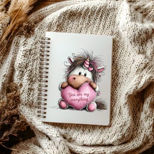 Puede incluir: Un cuaderno con espiral con una ilustraci&oacute;n de un unicornio sosteniendo un coraz&oacute;n rosa. El coraz&oacute;n tiene el texto "You are my everything". El unicornio tiene una melena marr&oacute;n y un cuerno rosa, sobre un fondo blanco.