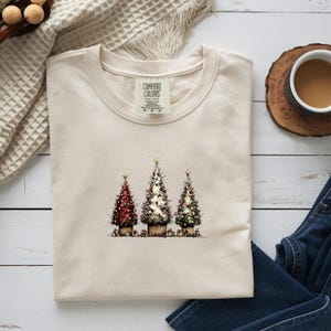 May include: A cream-colored t-shirt featuring a graphic print of three decorated Christmas trees in pots, with the Comfort Colors brand label visible at the neckline. The trees are adorned with lights and stars, set against a plain background.