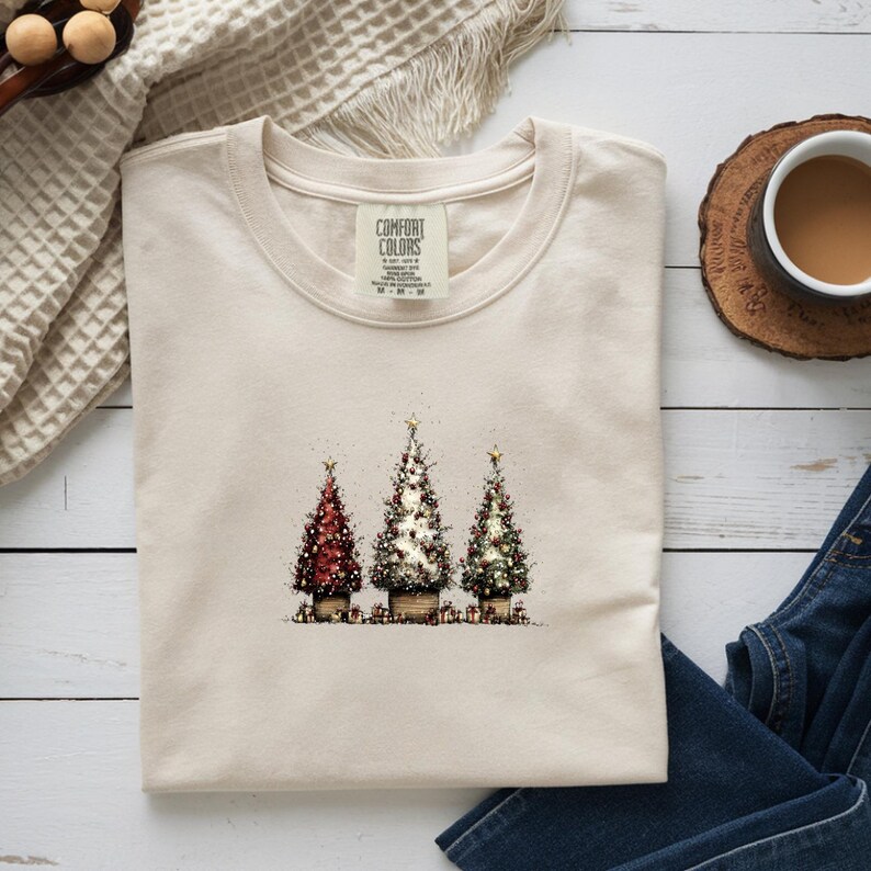 May include: A cream-colored t-shirt featuring a graphic print of three decorated Christmas trees in pots, with the Comfort Colors brand label visible at the neckline. The trees are adorned with lights and stars, set against a plain background.