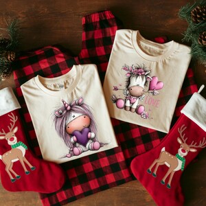 Puede incluir: Dos camisetas color crema con gr&aacute;ficos de unicornios, un pantal&oacute;n de pijama a cuadros rojos y negros y dos calcetines navide&ntilde;os rojos con dise&ntilde;os de renos. Una camiseta presenta un unicornio sosteniendo un coraz&oacute;n, la otra tiene un unicornio con una flecha en forma de coraz&oacute;n y la palabra "LOVE".