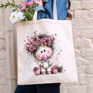 Puede incluir: Un bolso tote color crema con una ilustraci&oacute;n caprichosa de un unicornio. El unicornio tiene un anillo de diamantes rosa, una corona de flores y est&aacute; rodeado de corazones y flores rosas. El bolso lo lleva una persona con una chaqueta vaquera.