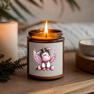 Puede incluir: Una vela encendida en un frasco de vidrio marr&oacute;n con una etiqueta que presenta un unicornio de dibujos animados con alas rosas. El unicornio tiene un coraz&oacute;n rosa. La vela est&aacute; en una bandeja de madera.