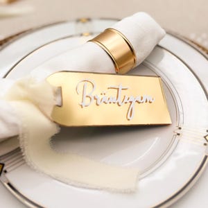 Könnte beinhalten: Ein Gedeck mit einer weißen Serviette, die von einem goldenen Ring gehalten wird, auf einem weißen Teller mit Goldrand. Ein goldenes Namensschild mit dem Wort "Bräutigam" ist mit einem cremefarbenen Band an der Serviette befestigt. Das Gedeck steht auf einem geflochtenen Platzset.