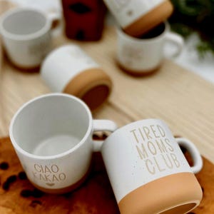 May include: White ceramic mugs with a speckled finish and tan-colored bases. One mug reads "CIAO KAKAO," another "TIRED MOMS CLUB," and others have text. The mugs are arranged on a wooden surface, suggesting a coffee or tea theme.