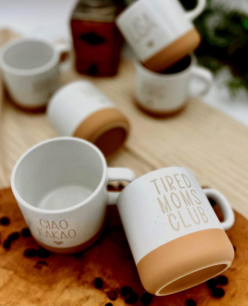 May include: White ceramic mugs with a speckled finish and tan-colored bases. One mug reads "CIAO KAKAO," another "TIRED MOMS CLUB," and others have text. The mugs are arranged on a wooden surface, suggesting a coffee or tea theme.