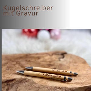 May include: Two wooden pens with silver accents and engraved text. One pen reads "Lehrerin mit" and the other reads "Kollegin mit", both with a heart symbol. The pens are displayed on a wooden surface.