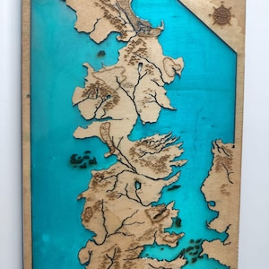 Puede incluir: Mapa de madera de Poniente con detalles cortados con láser de las masas terrestres y vías fluviales sobre un fondo turquesa. La palabra "PONIENTE" y una rosa de los vientos son visibles. Una pieza decorativa.