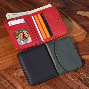 Puede incluir: Una cartera de cuero roja con cierre de botón a presión, abierta para revelar efectivo, tarjetas de crédito y una foto. También se ven una cartera negra y otra verde. Las carteras están sobre una superficie de madera.