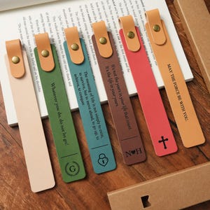 May include: A collection of colorful leather bookmarks with gold rivets and inspirational quotes. The bookmarks are in various colors, including beige, green, brown, and red. Each bookmark has a different quote printed on it.